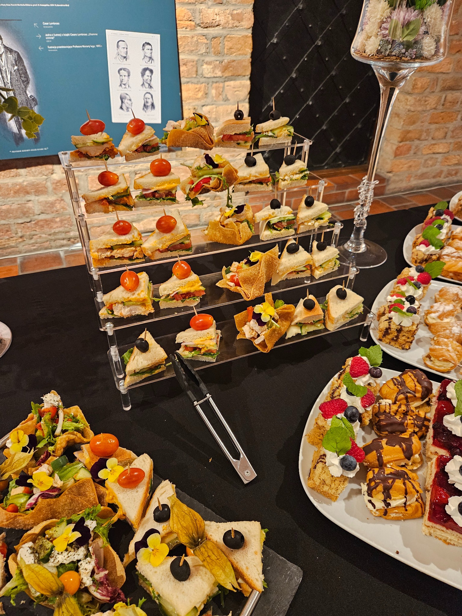 Elegancki stół cateringowy z różnorodnymi przekąskami: kanapkami na stojaku, tartaletkami i ciastami na talerzach. Dekoracje kwiatowe i ceglane tło dodają uroku.
