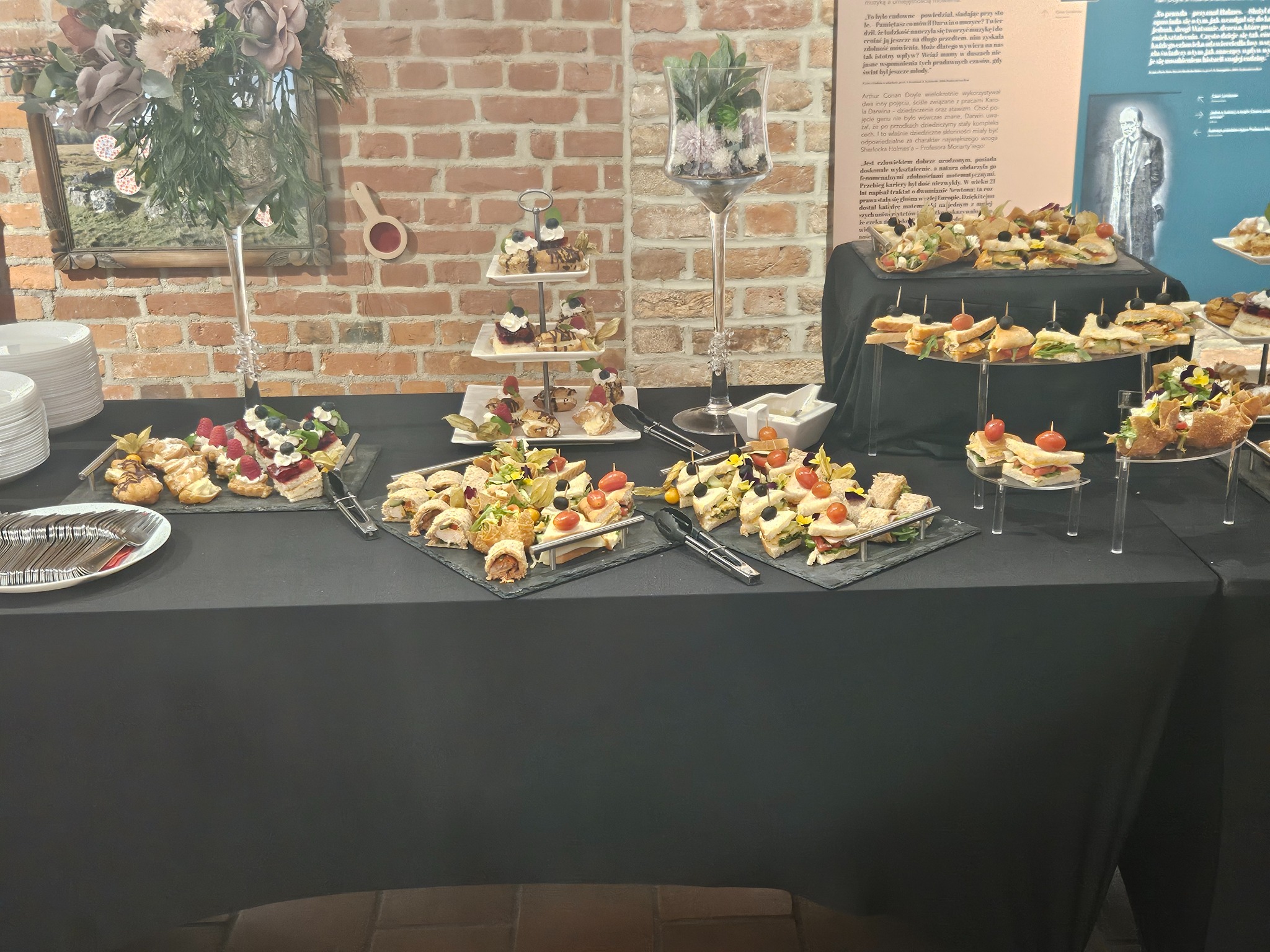 Stół cateringowy z przekąskami: mini kanapeczki, tartaletki z owocami, desery na paterze. Dekoracja kwiatowa i ceglana ściana w tle. Elegancki i apetyczny.