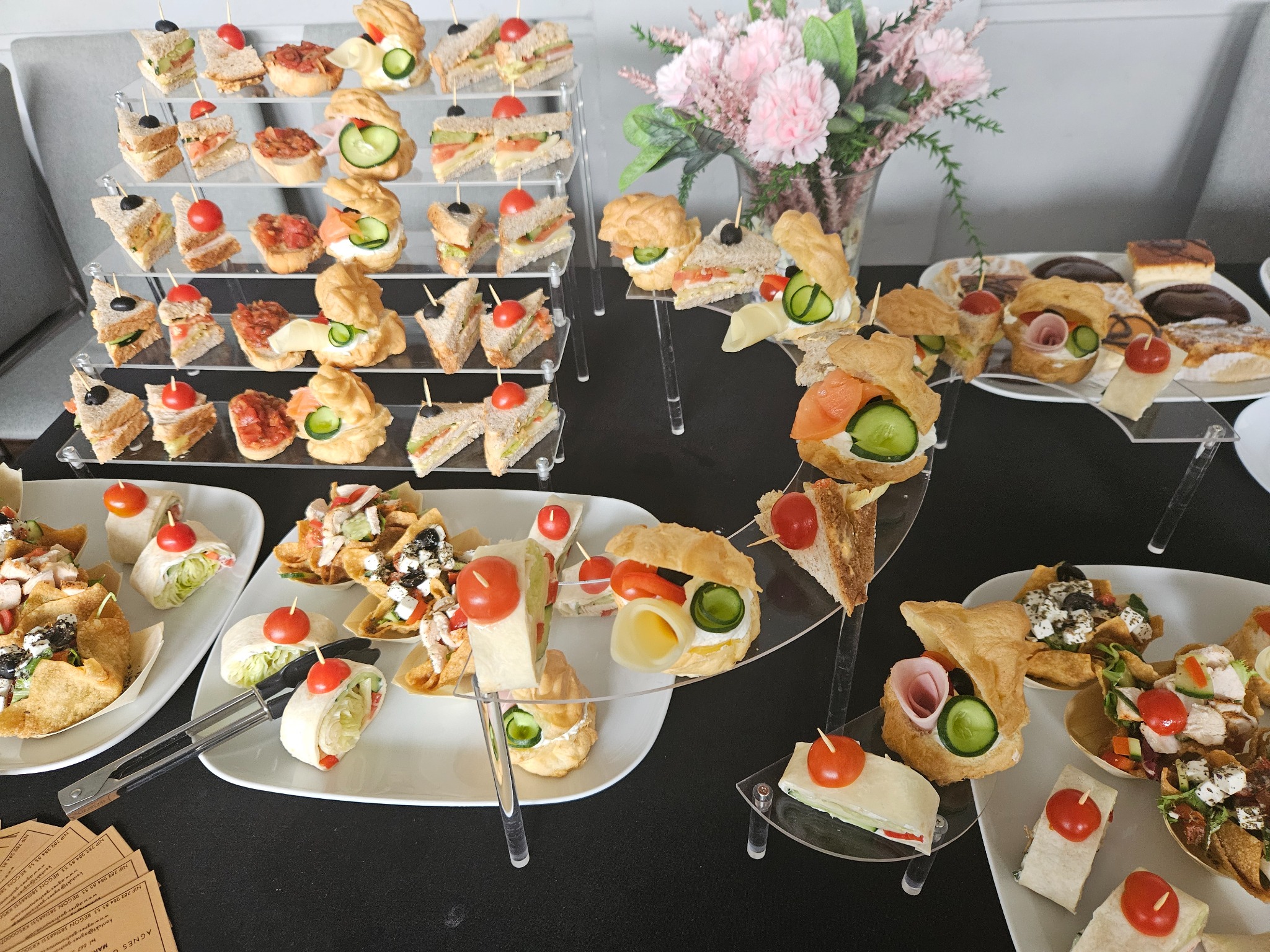 Elegancki catering: różnorodne przekąski na paterach i talerzach, w tym kanapki, tartaletki i roladki, udekorowane pomidorkami koktajlowymi, z kwiatami w tle.