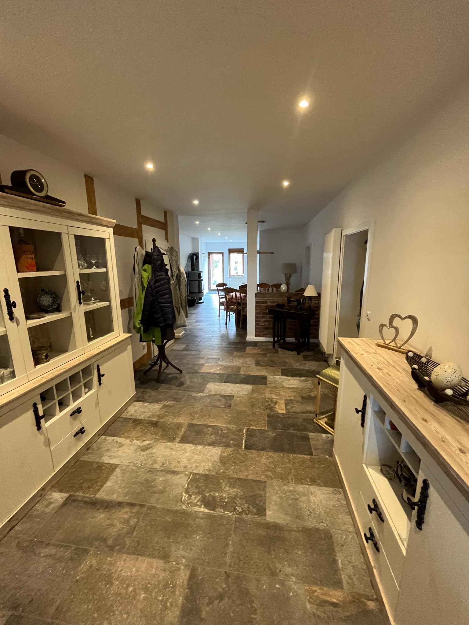 Wykończony korytarz domu z kamienną podłogą, białymi szafkami z ciemnymi uchwytami i drewnianymi elementami konstrukcyjnymi. W tle widoczny stół i krzesła.