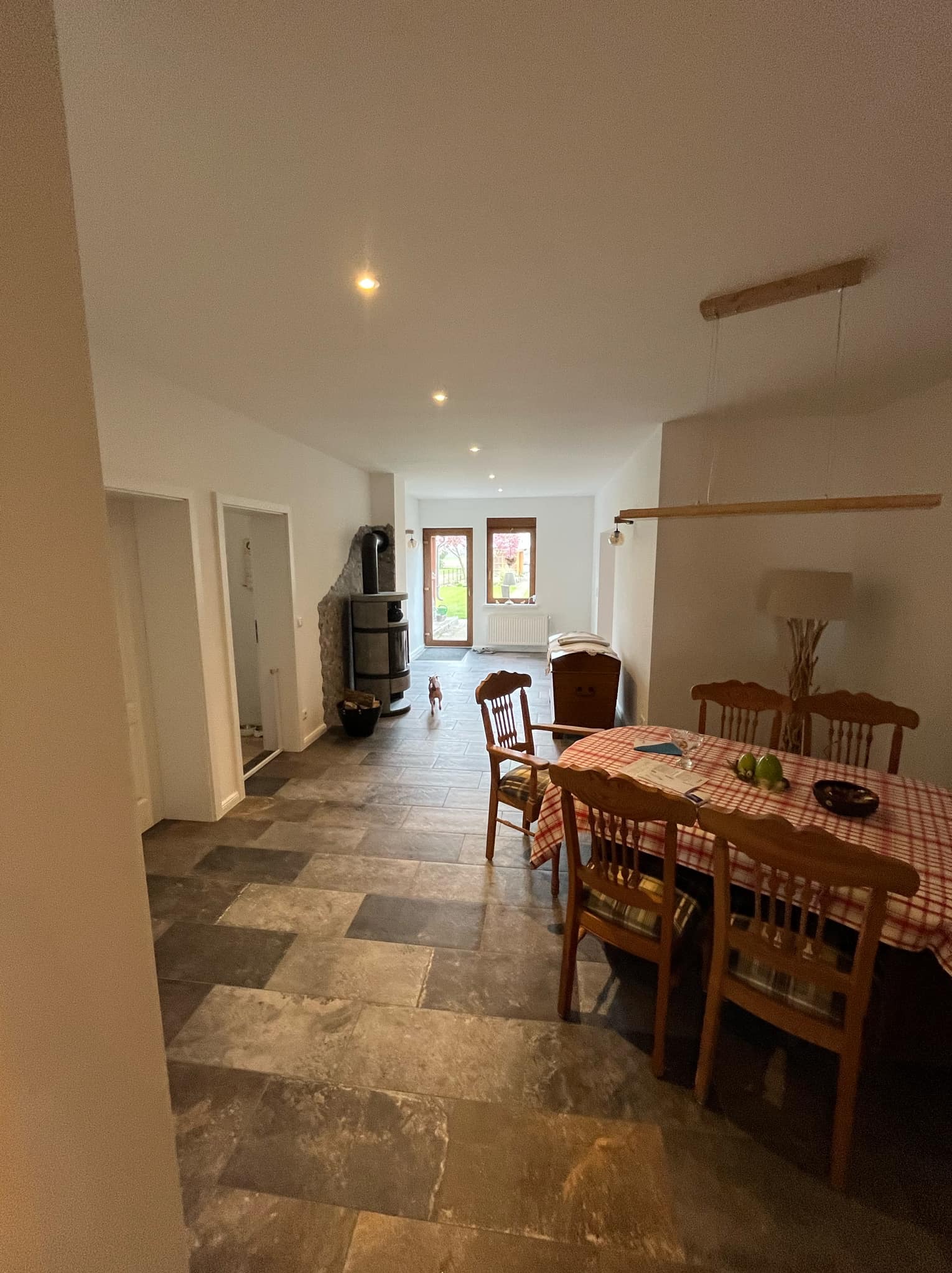 Jasne wnętrze z kamienną podłogą i drewnianym stołem. W tle piec kaflowy i okno z widokiem na ogród. Uwagę przykuwa pies rasy chihuahua.