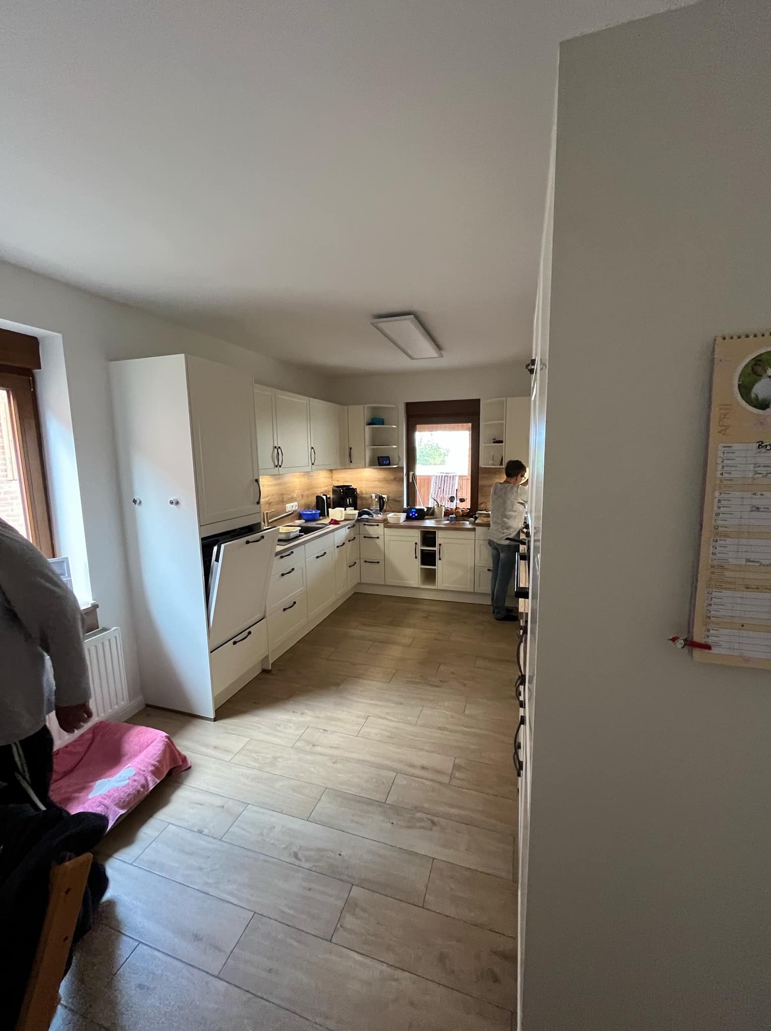 Jasna kuchnia w stylu skandynawskim z drewnianą podłogą i białymi meblami. Widoczna osoba przy zlewie i otwarte drzwi piekarnika.