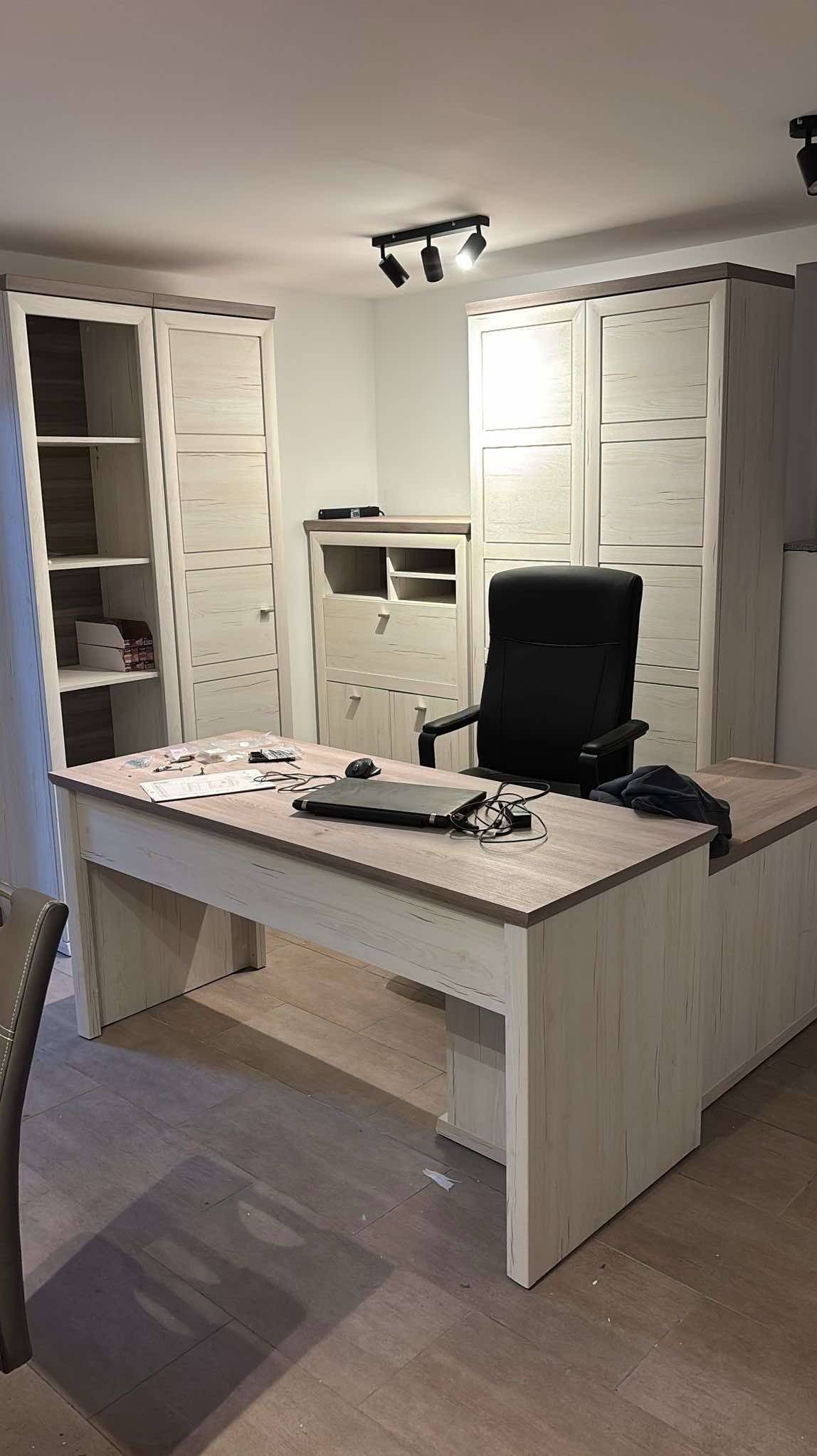 Nowoczesne biuro z drewnianym biurkiem, czarnym krzesłem i szafami w tle. Na biurku laptop i akcesoria biurowe. Oświetlenie punktowe na suficie. Jasne wnętrze.