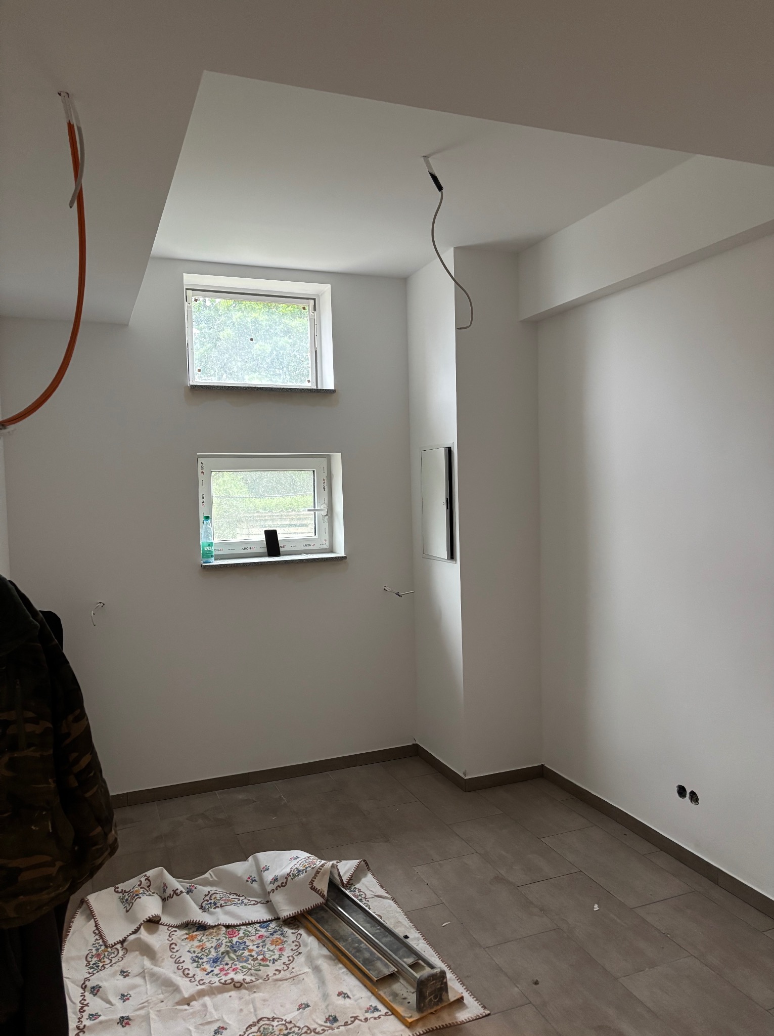 Wnętrze w trakcie wykańczania: białe ściany, dwa okna, brązowa podłoga z płytek. Widoczne narzędzia i materiały budowlane, sugerujące etap prac wykończeniowych.