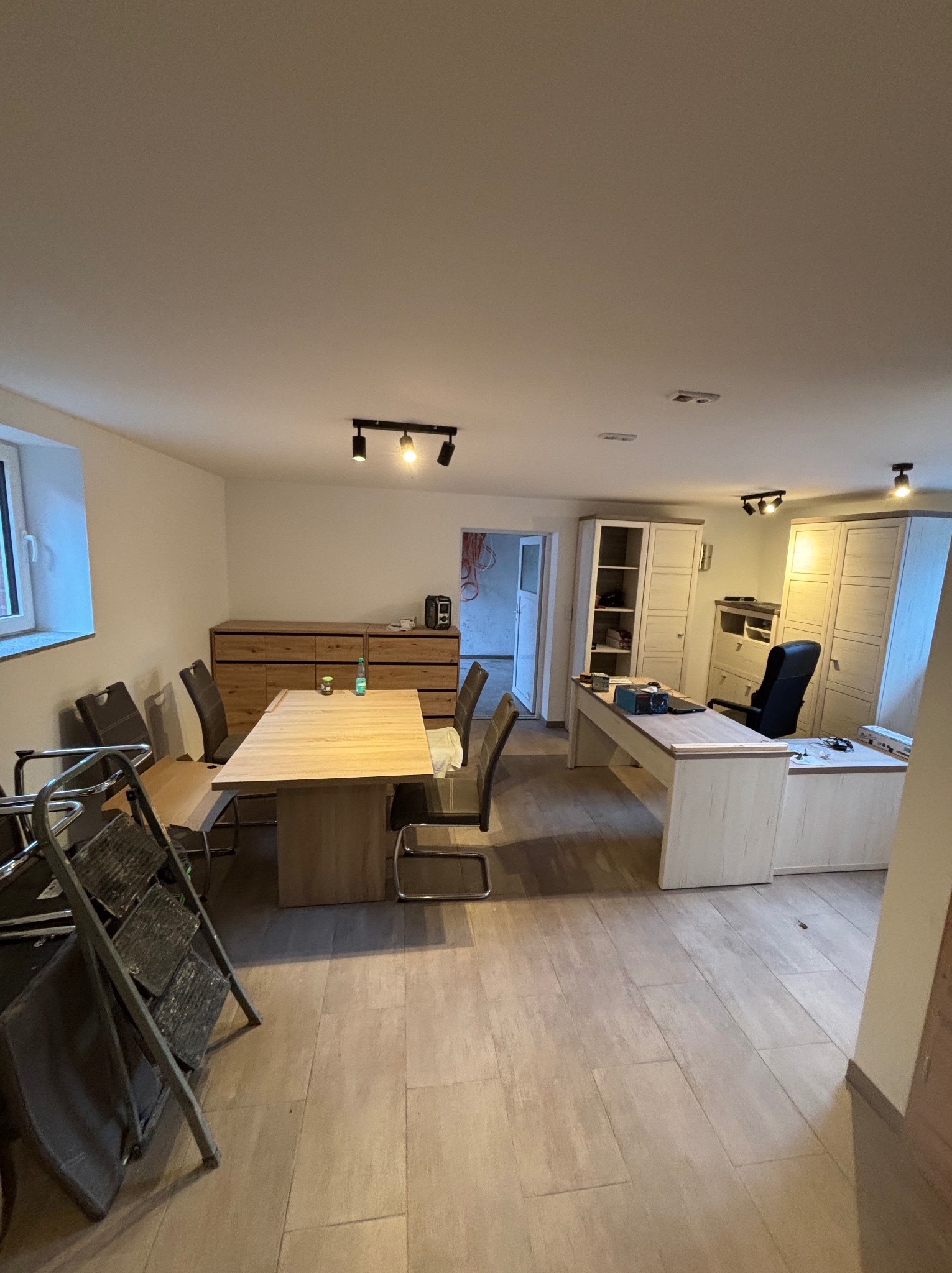 Jasne wnętrze po remoncie: stół jadalny, biurko z krzesłem biurowym, szafy. Na podłodze płytki imitujące drewno. W rogu drabina malarska.