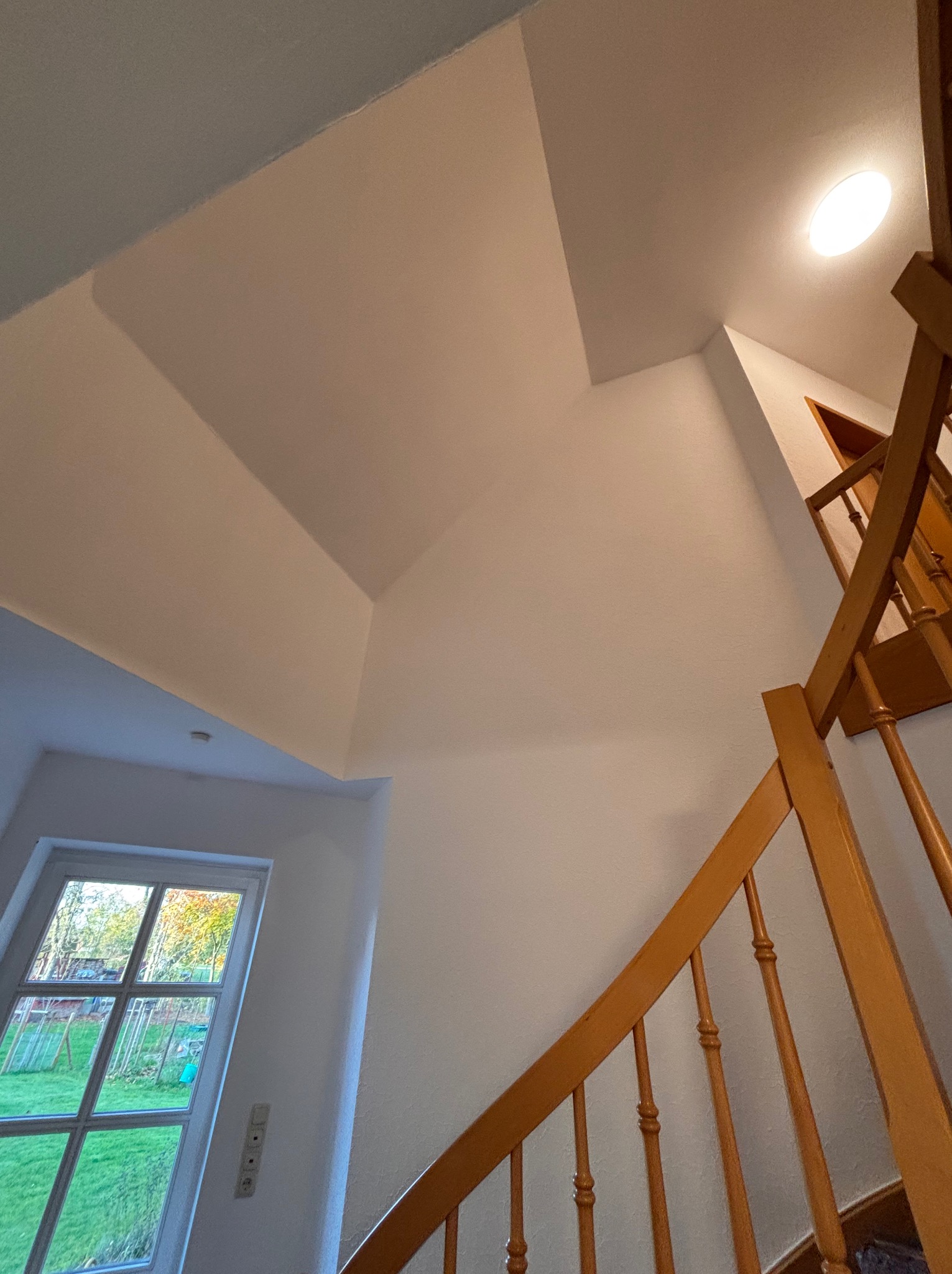 Jasne wnętrze z drewnianymi schodami i oknem. Ściany pomalowane na biało, sufit z geometrycznymi wnękami. Widok z dołu, podkreślający wysokość pomieszczenia.