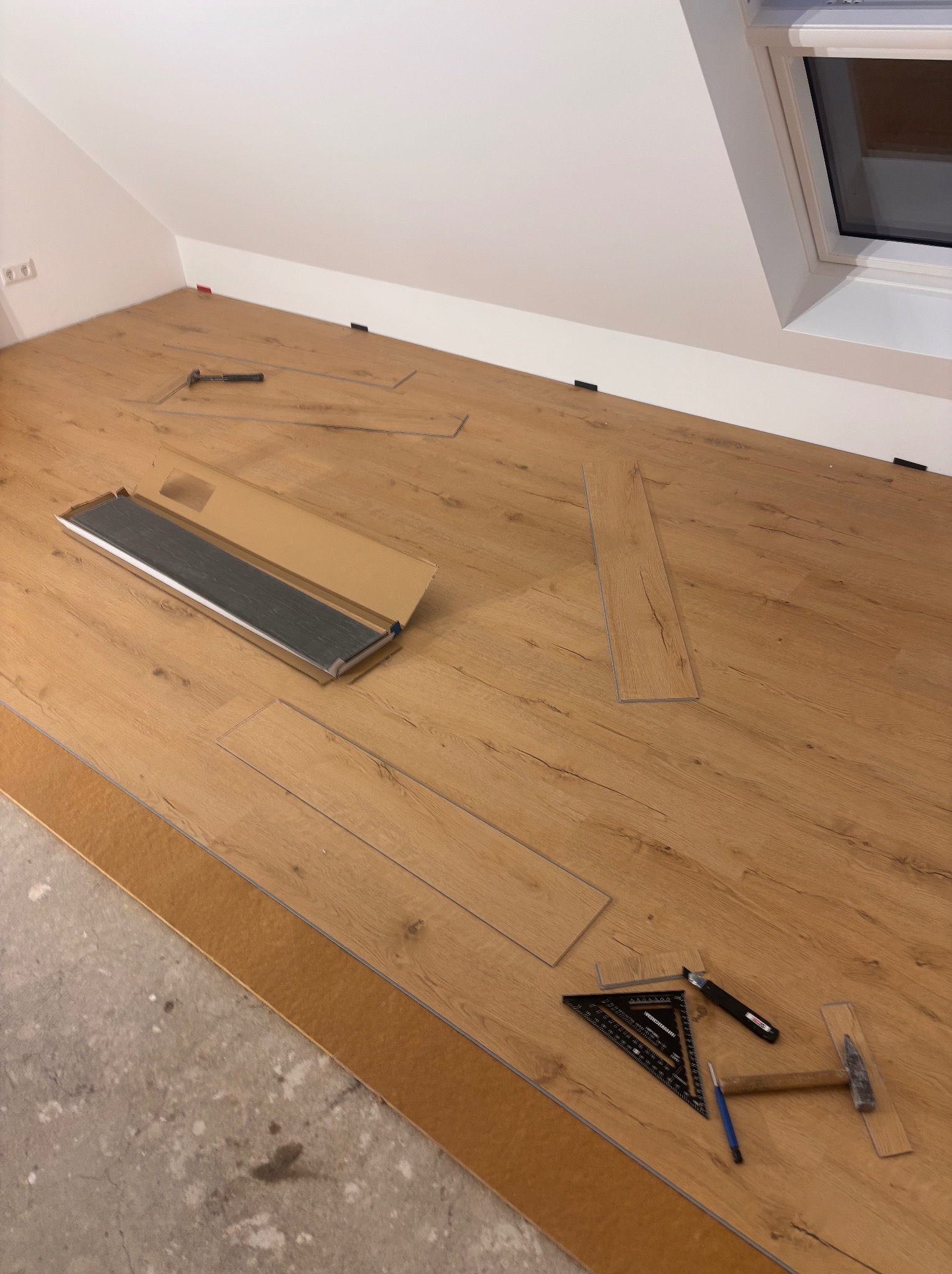 Montaż paneli podłogowych w pokoju na poddaszu. Widoczne narzędzia: młotek, kątownik, ołówek. Część podłogi wyłożona, reszta w trakcie prac. Okno dachowe w tle.