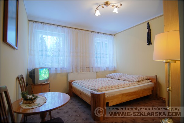 Pokój gościnny w Szklarskiej Porębie z drewnianym łóżkiem, telewizorem i małym stolikiem z dekoracją kwiatową.