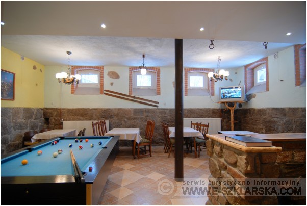 Wnętrze pokoi gościnnych z kamienną podmurówką, stołami nakrytymi białymi obrusami, stołem bilardowym i telewizorem na drewnianym stelażu.