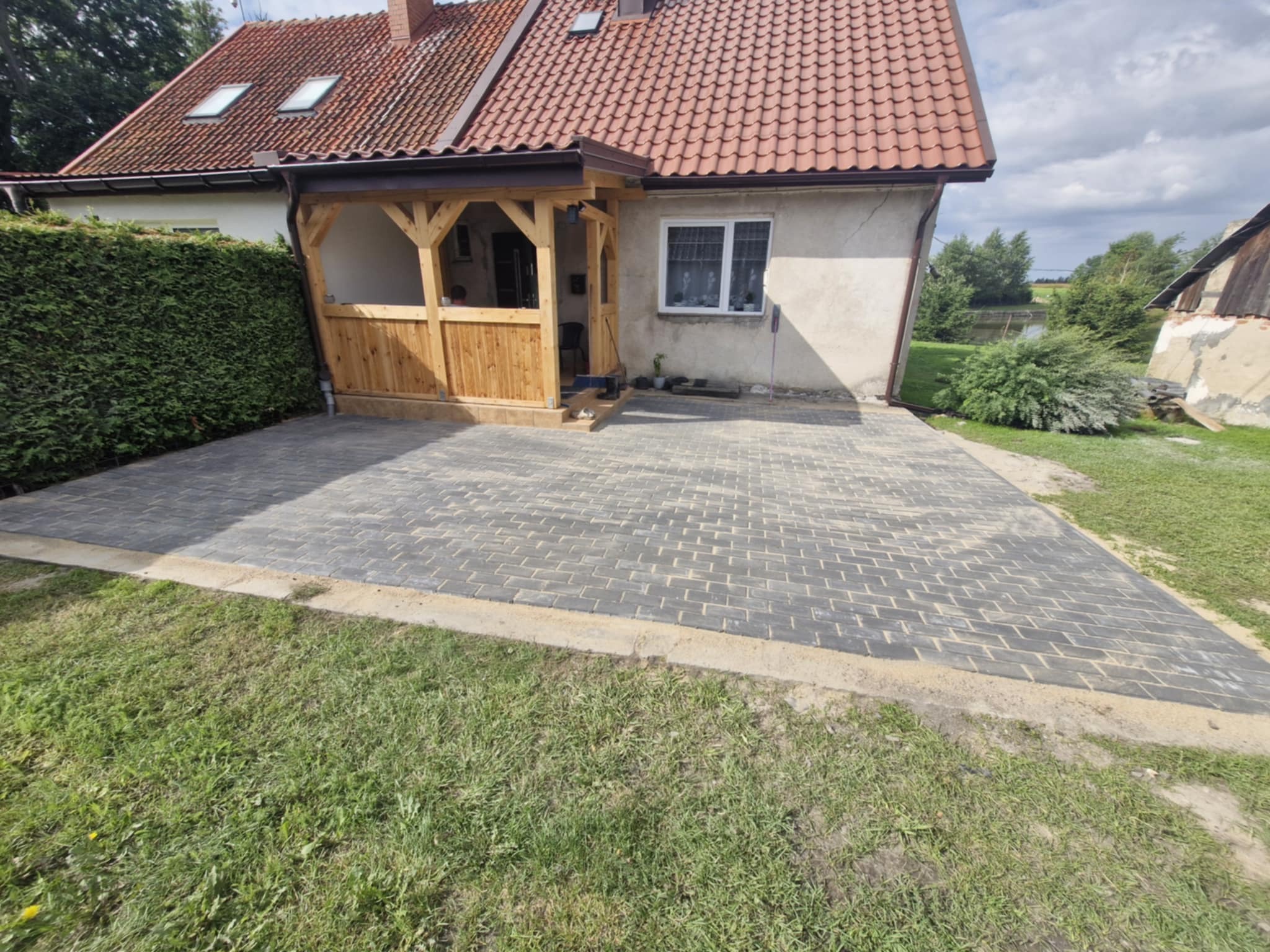 Ułożona, szara kostka brukowa przed domem z drewnianą werandą. Widoczne zielone trawniki i krzewy. Całość w słoneczny dzień.