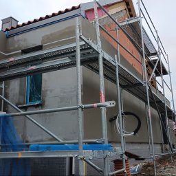 AStat-Bud - Budynek w trakcie prac elewacyjnych, widoczne rusztowania, ocieplenie i nałożony tynk. Częściowo otynkowana ściana, okno zabezpieczone folią. Prace na zewnątrz budynku.