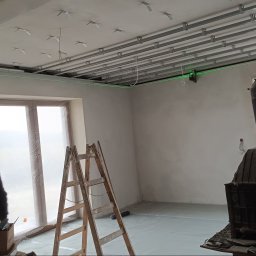 AStat-Bud - Wnętrze w trakcie prac: widoczny stelaż sufitu podwieszanego z profilami, poziomica laserowa, drabina i piec typu koza. Okno zabezpieczone folią.