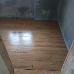 AStat-Bud - Ułożona podłoga z płytek imitujących drewno w surowym wnętrzu łazienki. Widoczne przygotowane ściany i fragmenty niedokończonej podłogi przy wejściu.