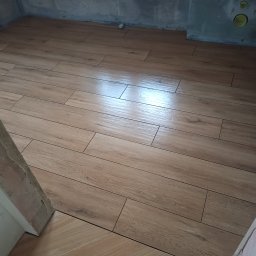 AStat-Bud - Ułożona podłoga z płytek imitujących drewno w surowym pomieszczeniu. Widoczne niedokończone ściany i fragmenty futryny. Płytki ułożone w regularny wzór.