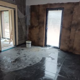 AStat-Bud - Remont łazienki w trakcie: położone brązowe płytki na ścianach, czarne płytki na podłodze z połyskiem, zabezpieczone okno folią, widoczne ślady prac i wiadro farby.