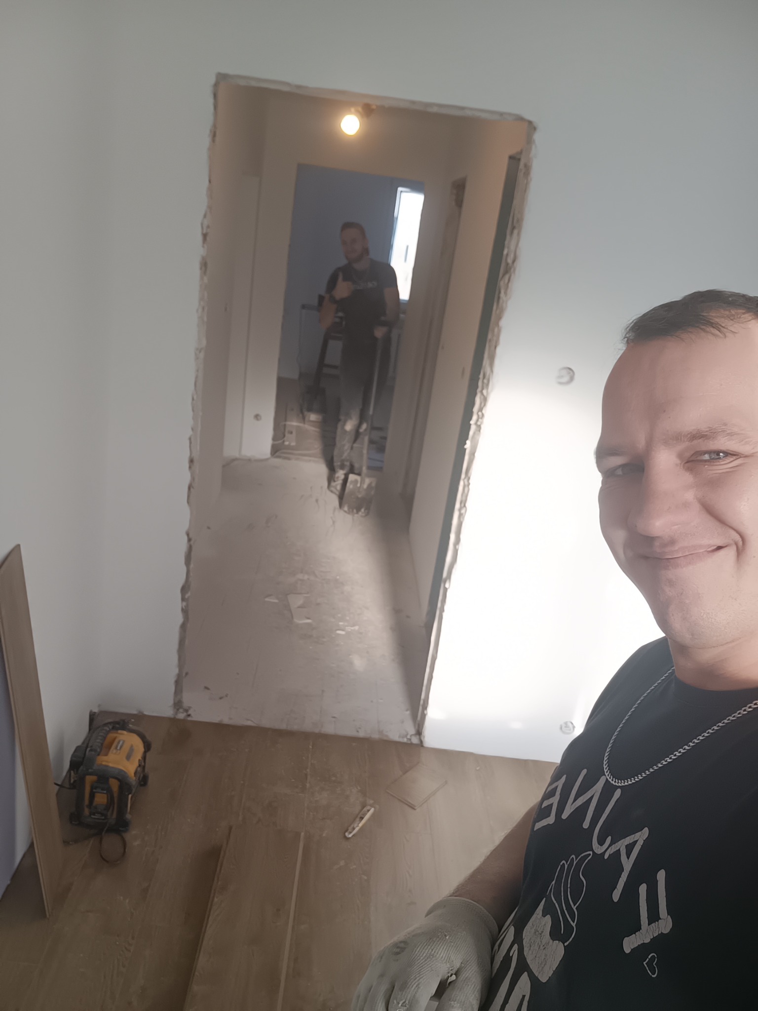 Układanie parkietu w trakcie remontu mieszkania. Widoczny pracownik z uśmiechem, narzędzia i częściowo ułożona podłoga. W tle drugi pracownik w przejściu.