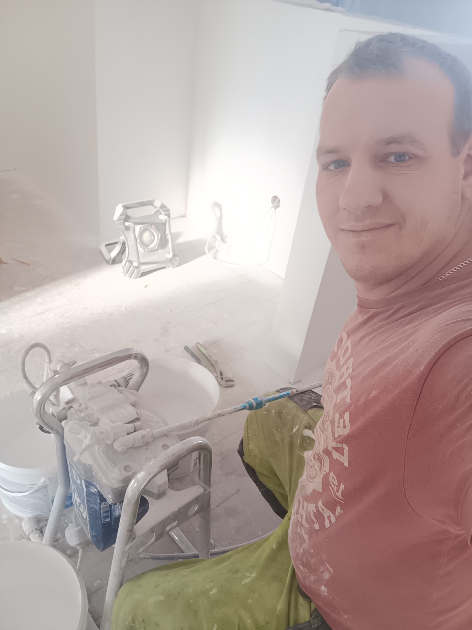 Uśmiechnięty malarz z urządzeniem do natryskowego malowania ścian w jasnym wnętrzu. Widoczne ślady farby na ubraniu i sprzęcie.