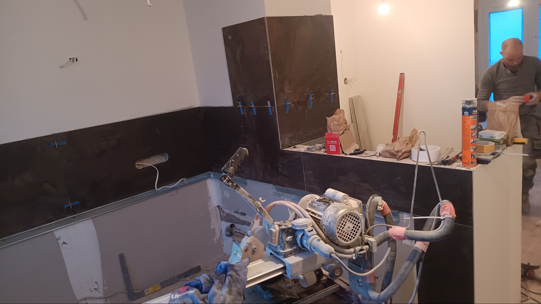 Częściowo wyłożona czarną płytką ściana łazienki z widoczną maszyną do cięcia płytek i pracującym glazurnikiem w tle. Nieukończone wykończenie.