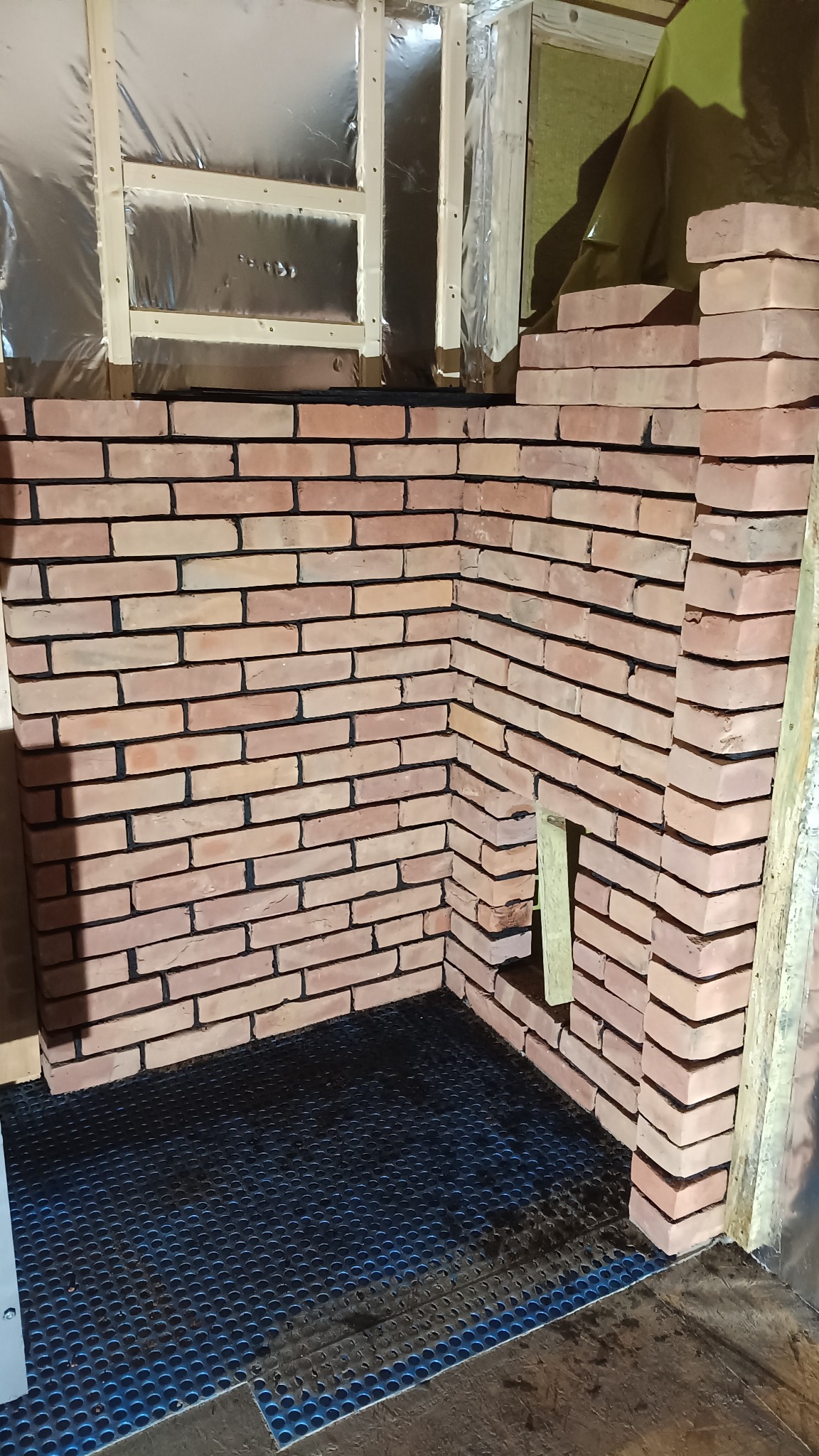 Wnętrze w trakcie prac murarskich: ściana z czerwonej cegły, widoczne izolacje i podłoże z perforowanej gumy. Etap budowy lub wykończenia z bliska.