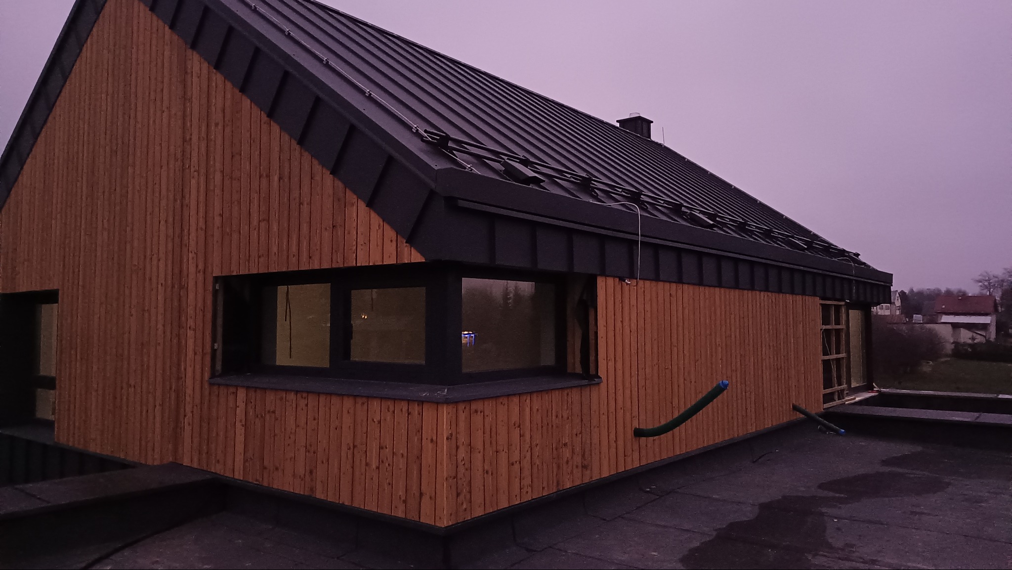 Nowoczesny dom z drewnianą elewacją i czarnym dachem z blachy na tle pochmurnego nieba. Widoczne okna z czarnymi ramami i fragment dachu pokrytego papą.