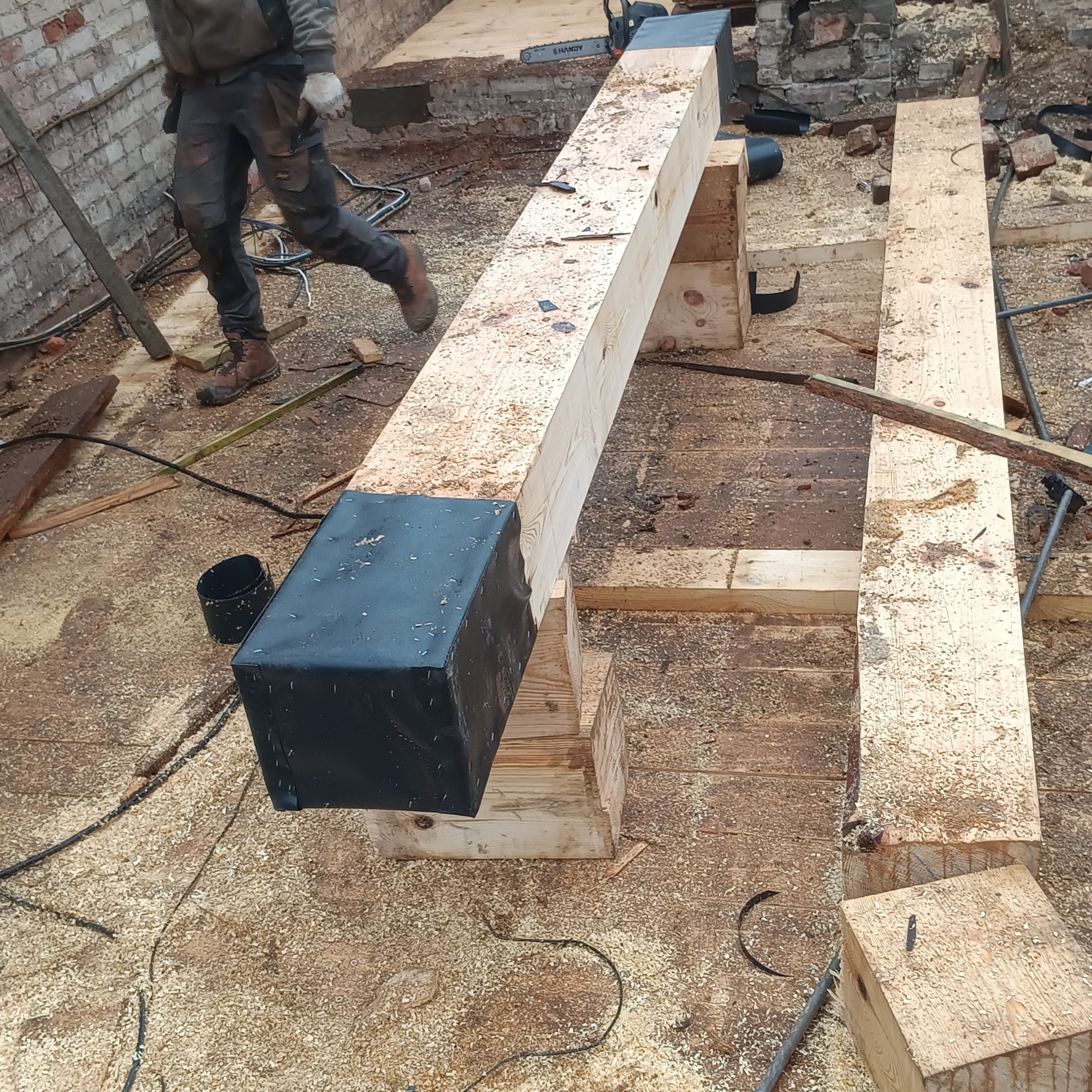 Drewniana belka wzmocniona czarną folią, podparta na drewnianych klockach. W tle widać mur z cegły i osobę w roboczym ubraniu. Wszędzie wióry.