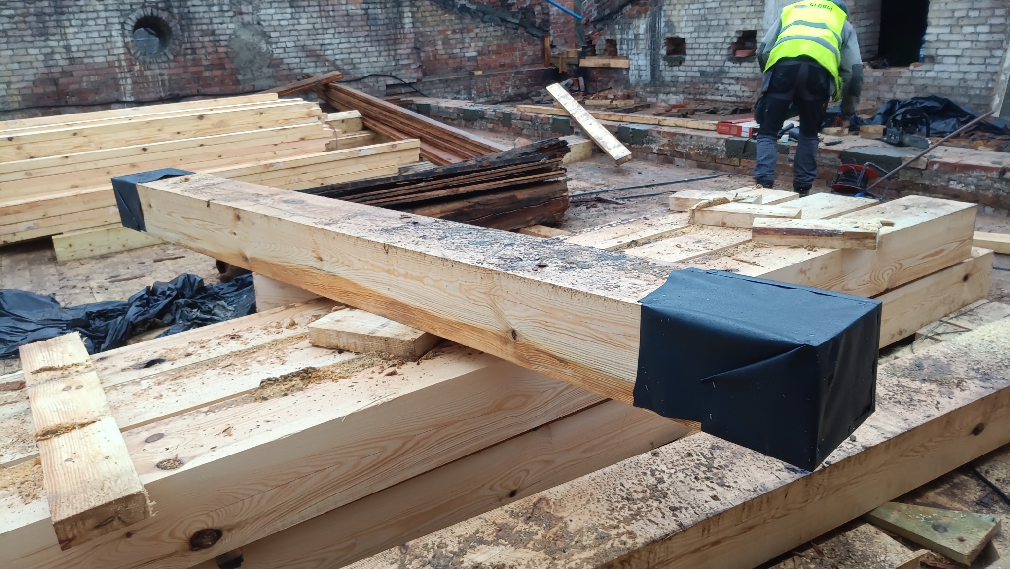 Montaż drewnianej konstrukcji stropu z belek, zabezpieczonych czarną folią. W tle mur z cegły i pracownik w kamizelce odblaskowej. Widoczne ślady obróbki drewna.