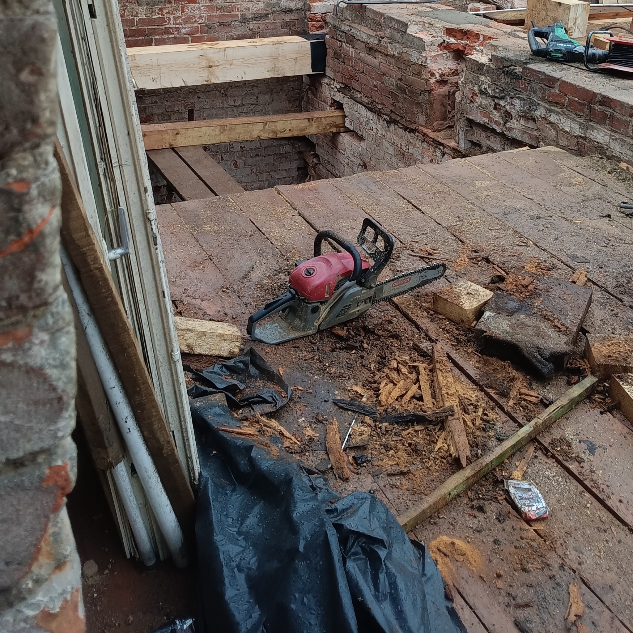 Demontaż drewnianej podłogi na balkonie z użyciem piły łańcuchowej. Widoczne ceglane elementy konstrukcyjne i pozostałości po pracach rozbiórkowych.