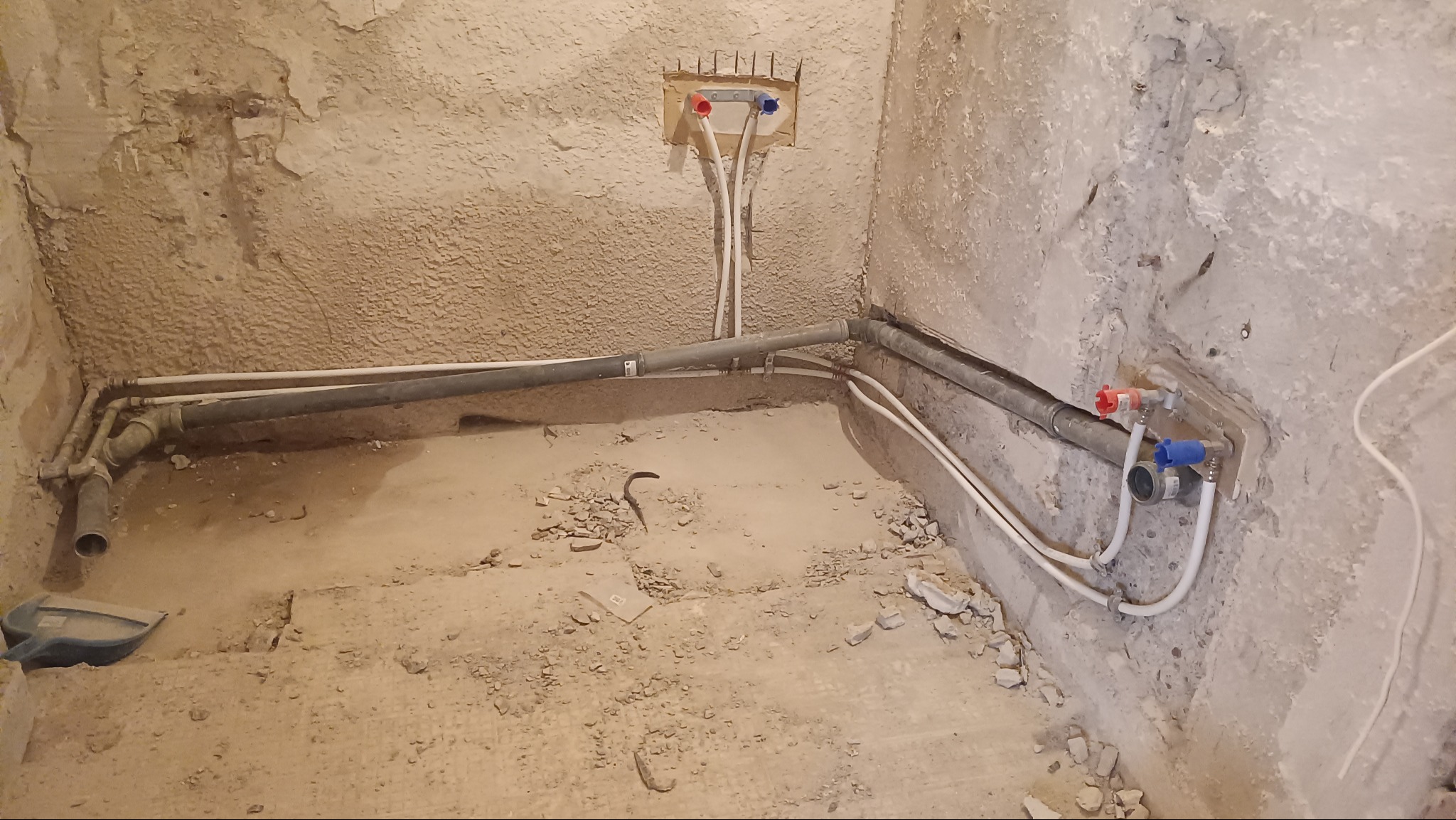 Surowy stan łazienki w trakcie prac: widoczne rury wodne i kanalizacyjne, ściany w stanie deweloperskim, podłoga z resztkami gruzu. Instalacja wod-kan w trakcie realizacji.