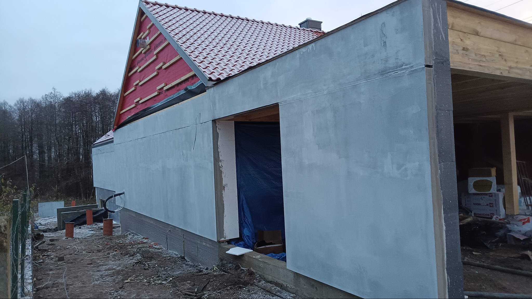 Budynek w trakcie szpachlowania zewnętrznego, widoczne fragmenty dachu z czerwoną dachówką i przygotowane otwory okienne zabezpieczone folią. Prace wykończeniowe.