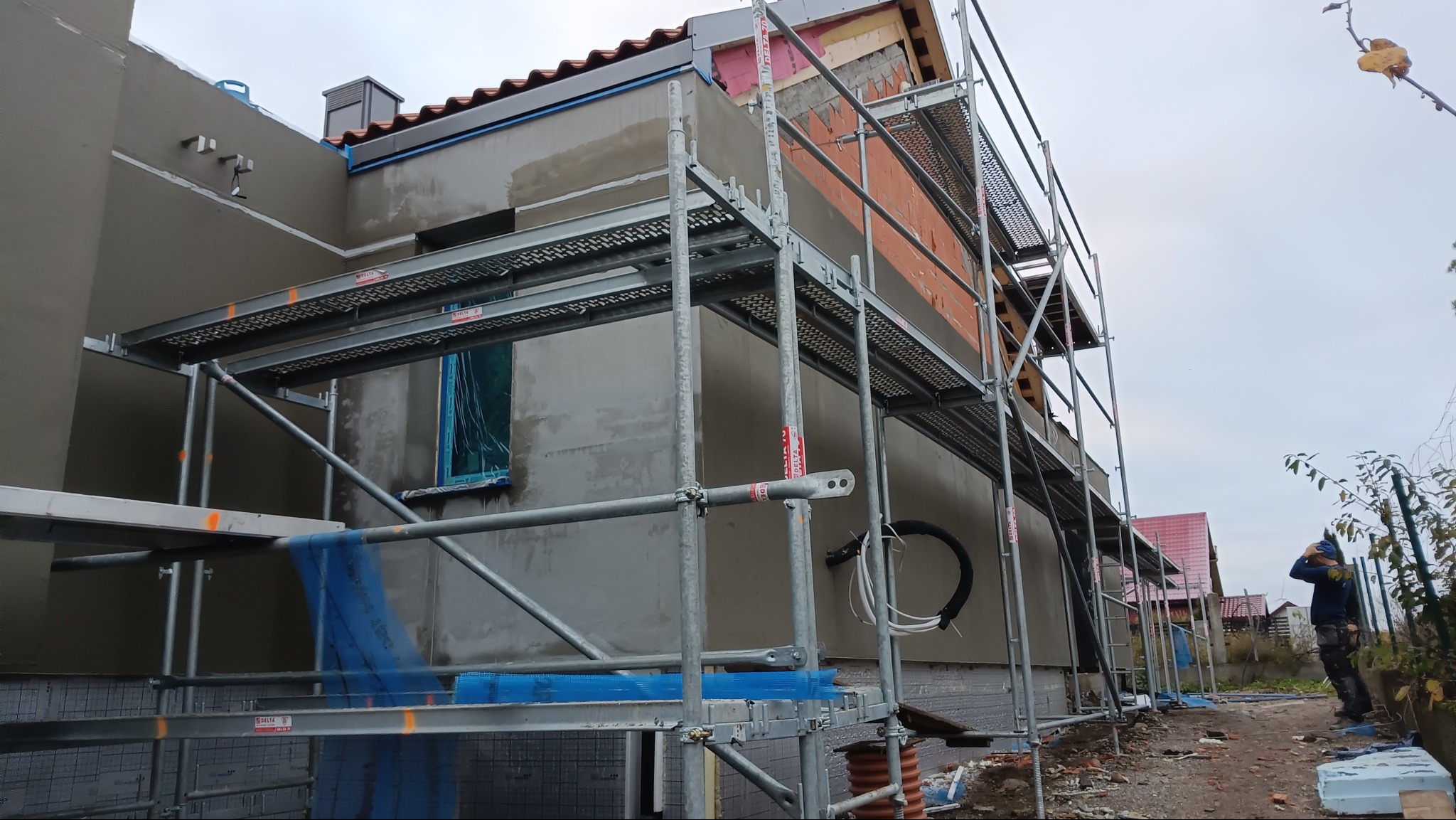 Budynek w trakcie prac elewacyjnych, widoczne rusztowania, ocieplenie i nałożony tynk. Częściowo otynkowana ściana, okno zabezpieczone folią. Prace na zewnątrz budynku.