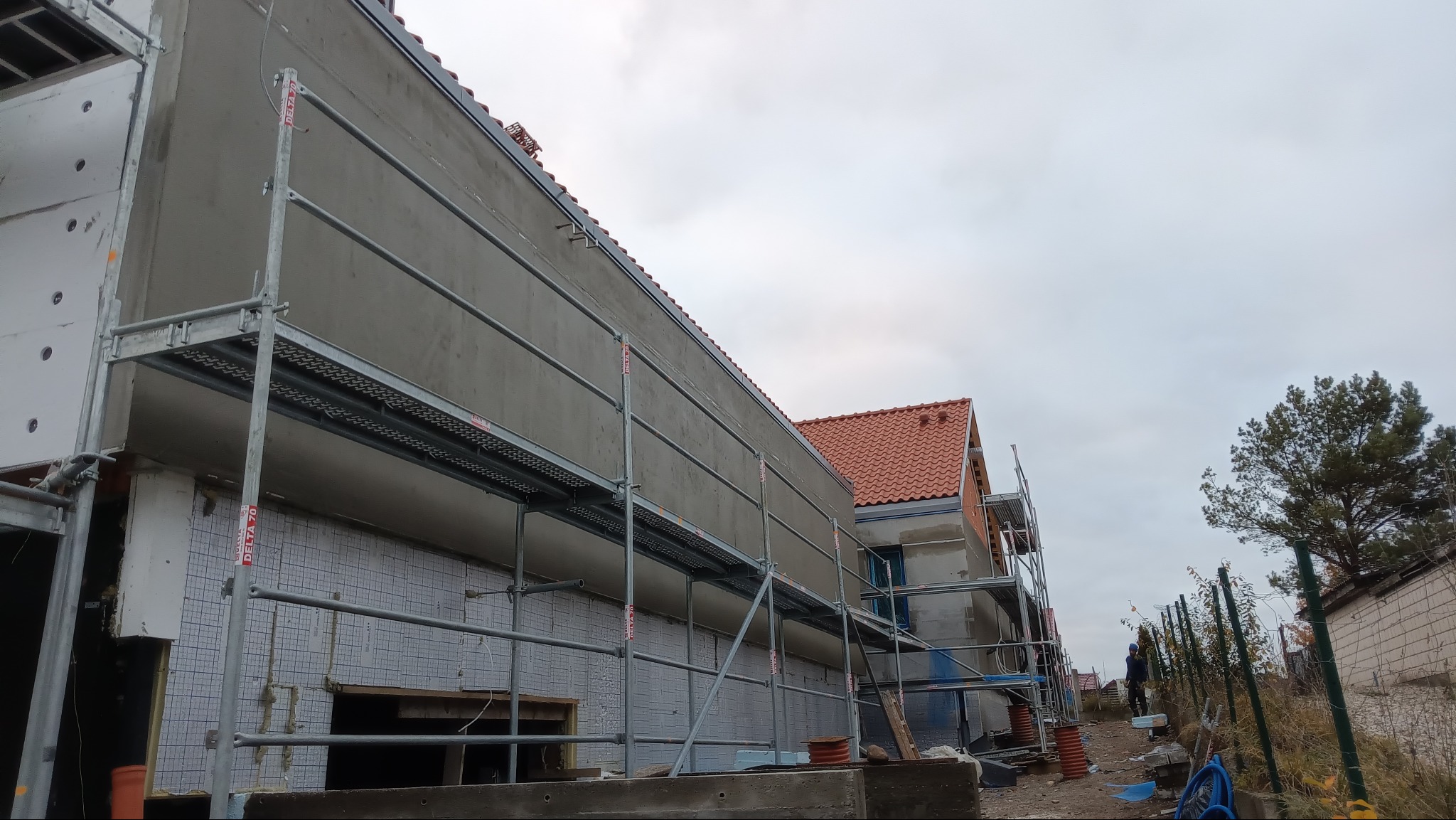 Budynek w trakcie ocieplania, widoczne rusztowania i warstwy izolacyjne. Szara elewacja kontrastuje z czerwoną dachówką. Prace w toku, widoczny pracownik w oddali.