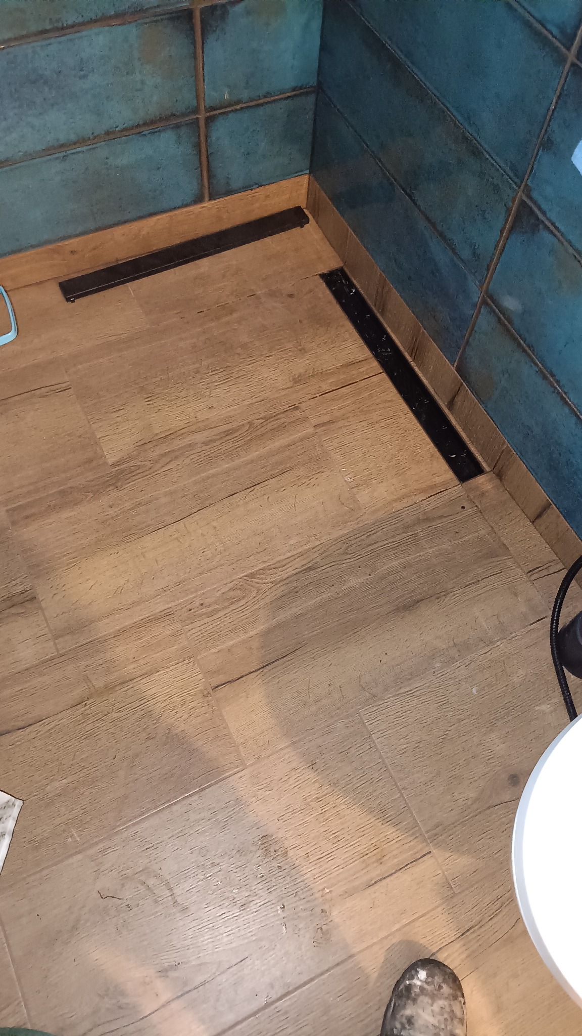 Wykończona łazienka z drewnopodobną podłogą i turkusowymi płytkami. Widoczne czarne odpływy liniowe przy ścianach. Ujęcie z góry, fragment buta w rogu kadru.