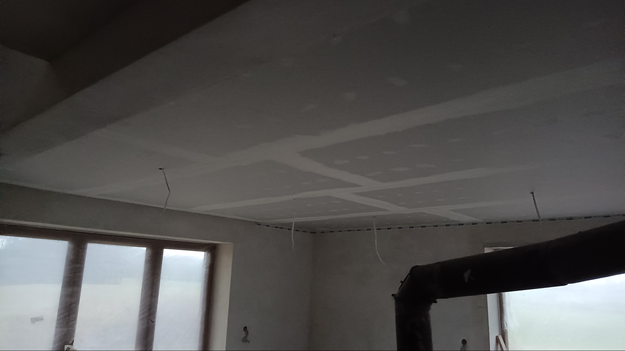 Sufit podwieszany z płyt gipsowo-kartonowych w trakcie montażu, widoczne łączenia i wystające przewody. W tle okno z widokiem na zewnątrz i fragment rury pieca.