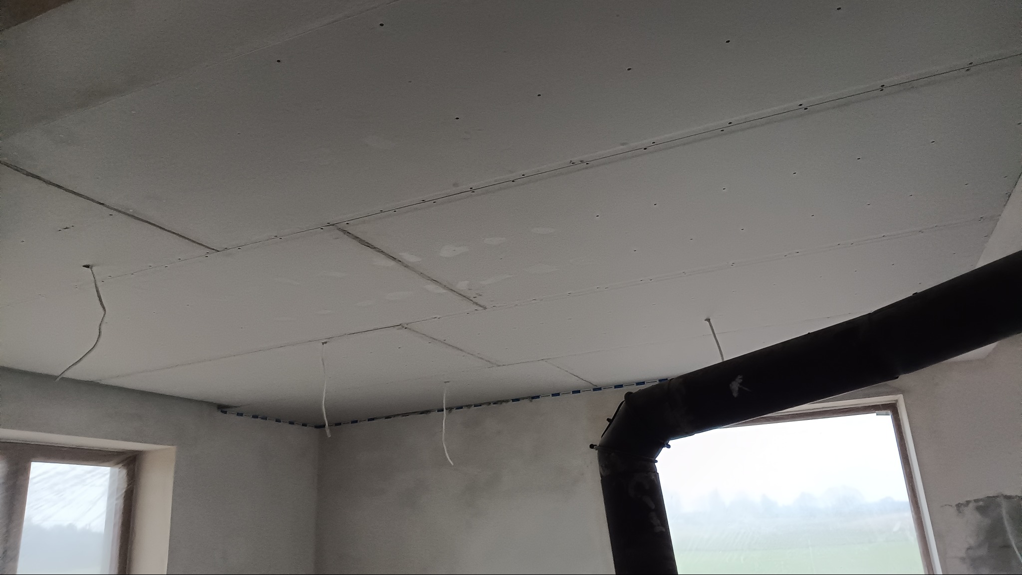 Sufit podwieszany z płyt gipsowo-kartonowych w trakcie montażu, widoczne łączenia płyt i przewody. Fragment okna i rury w tle. Surowy stan wykończenia.