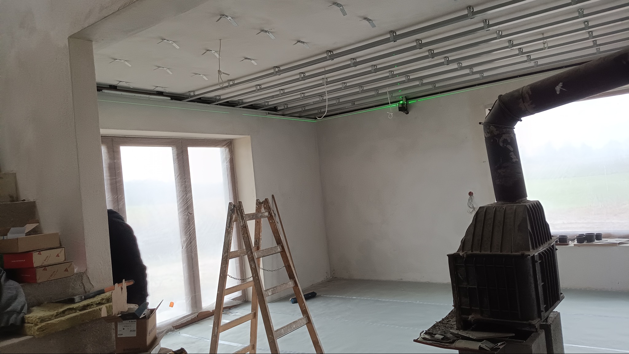 Wnętrze w trakcie prac: widoczny stelaż sufitu podwieszanego z profilami, poziomica laserowa, drabina i piec typu koza. Okno zabezpieczone folią.