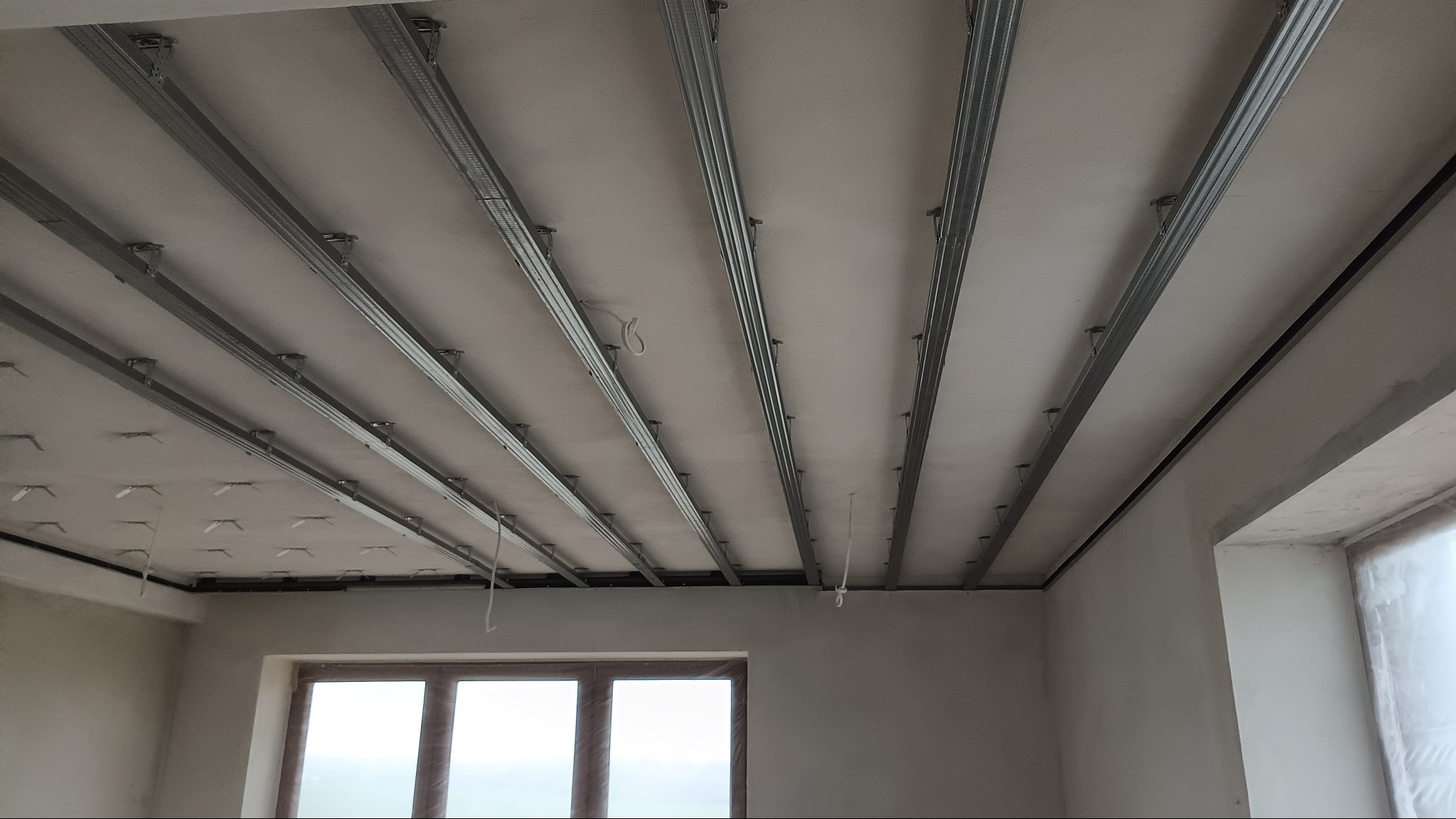 Montaż metalowego stelażu sufitu podwieszanego w Kolonii Sejny. Widoczne profile i mocowania, przygotowane do dalszych prac wykończeniowych.
