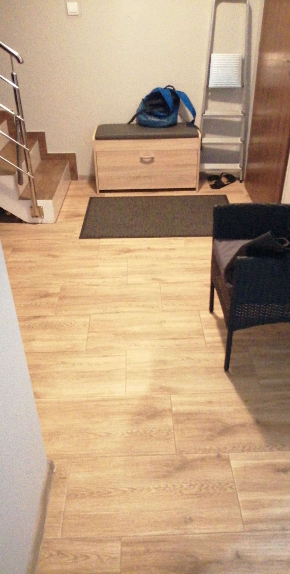 Jasne panele podłogowe imitujące drewno, ułożone w korytarzu. Widoczny fragment schodów, ławka z siedziskiem, drabina i krzesło z ciemnej wikliny.