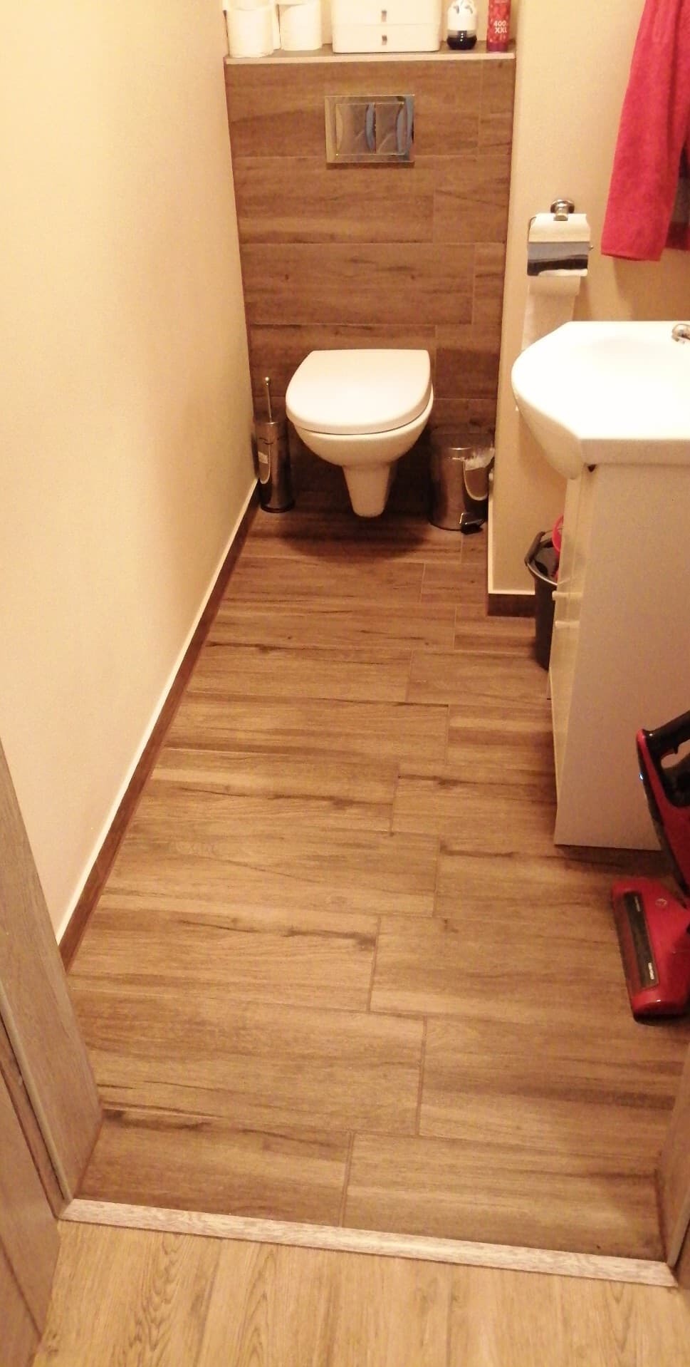 Ukończona łazienka z drewnopodobnymi płytkami na podłodze i ścianie za toaletą. Widoczna toaleta, umywalka i akcesoria łazienkowe.
