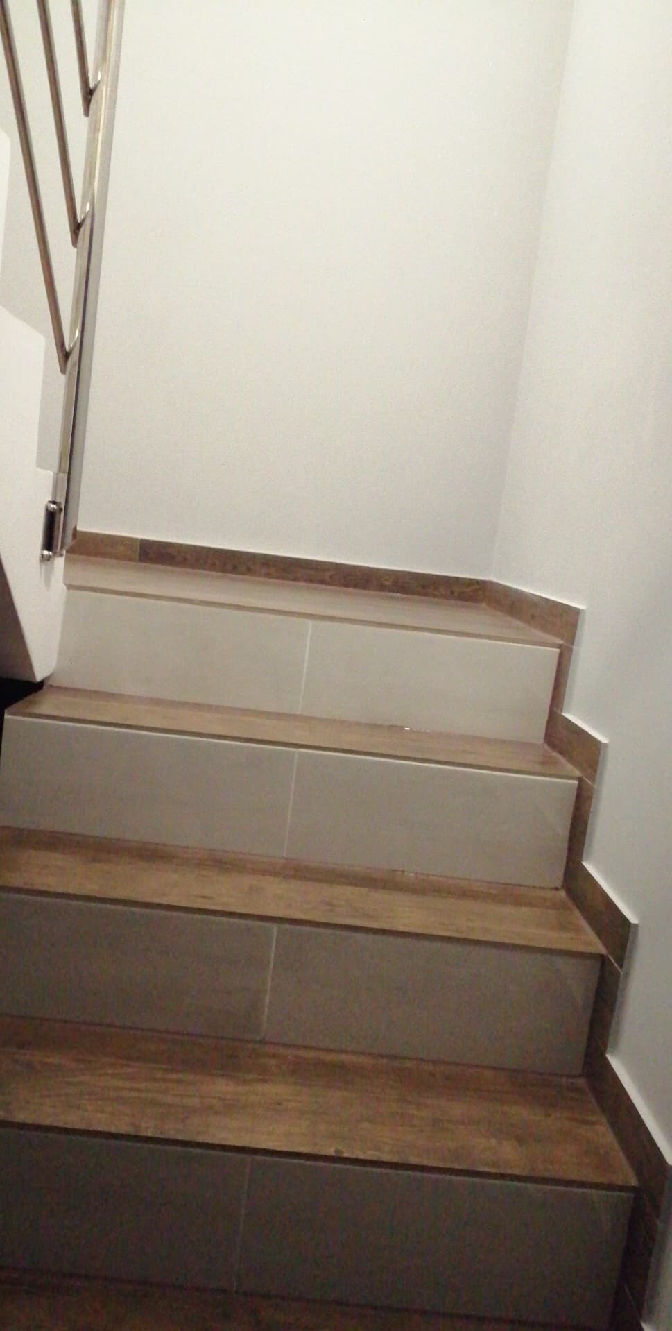 Wykończone schody z płytek imitujących drewno, z metalową barierką. Widok z dołu, ukazujący detale wykończenia i połączenia płytek.