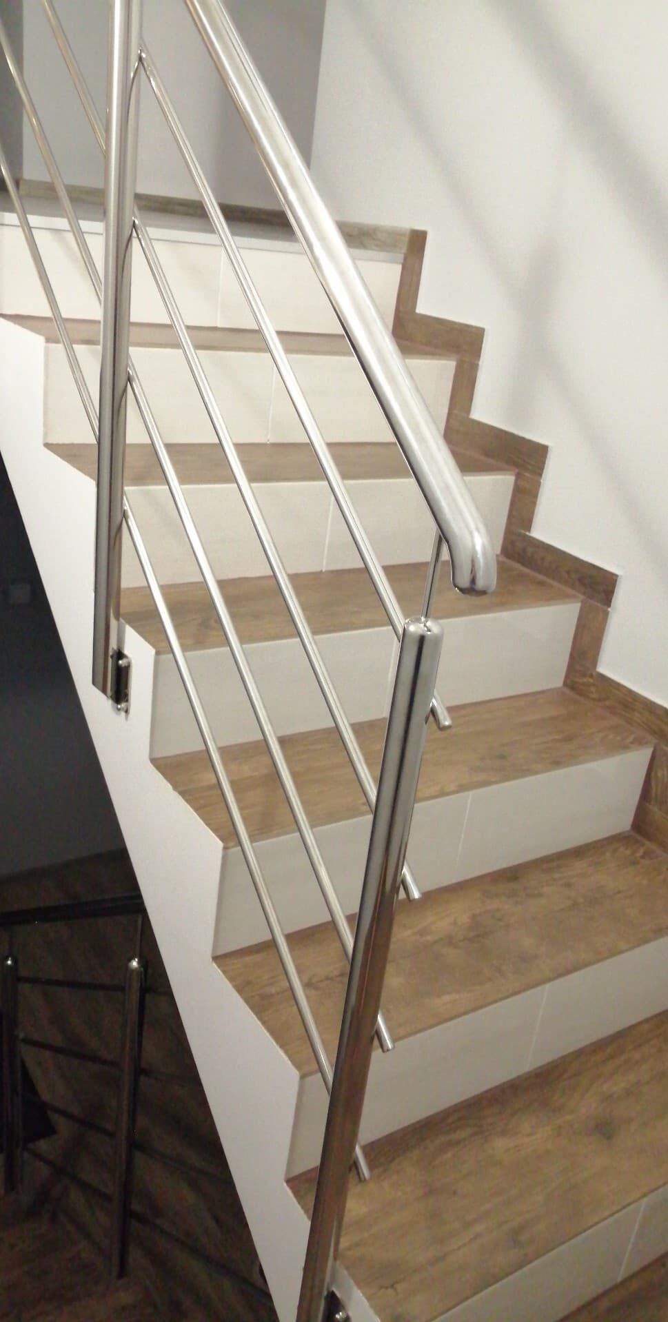 Schody z drewnopodobnymi płytkami i metalową balustradą. Widoczne detale wykończenia stopni i nowoczesny design poręczy.