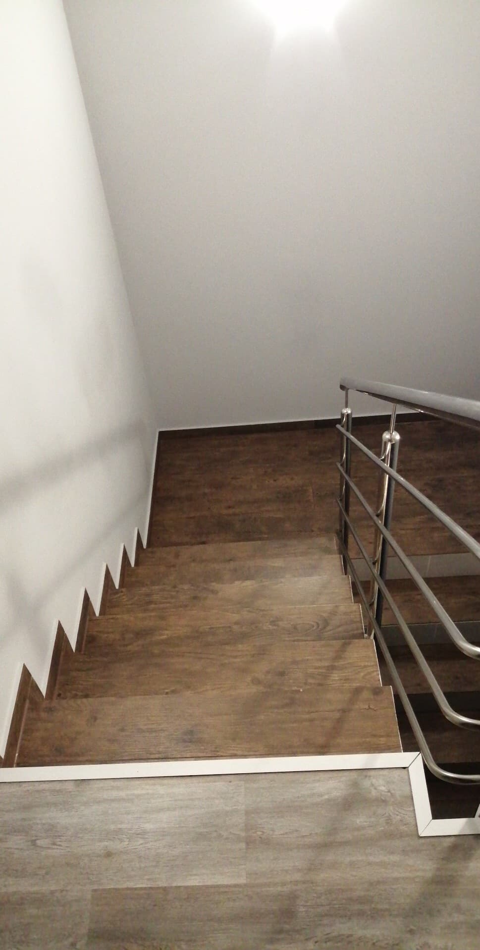 Schody wykończone panelami imitującymi drewno, z metalową balustradą. Widok z góry, oświetlenie sufitowe. Białe ściany i listwy przypodłogowe.