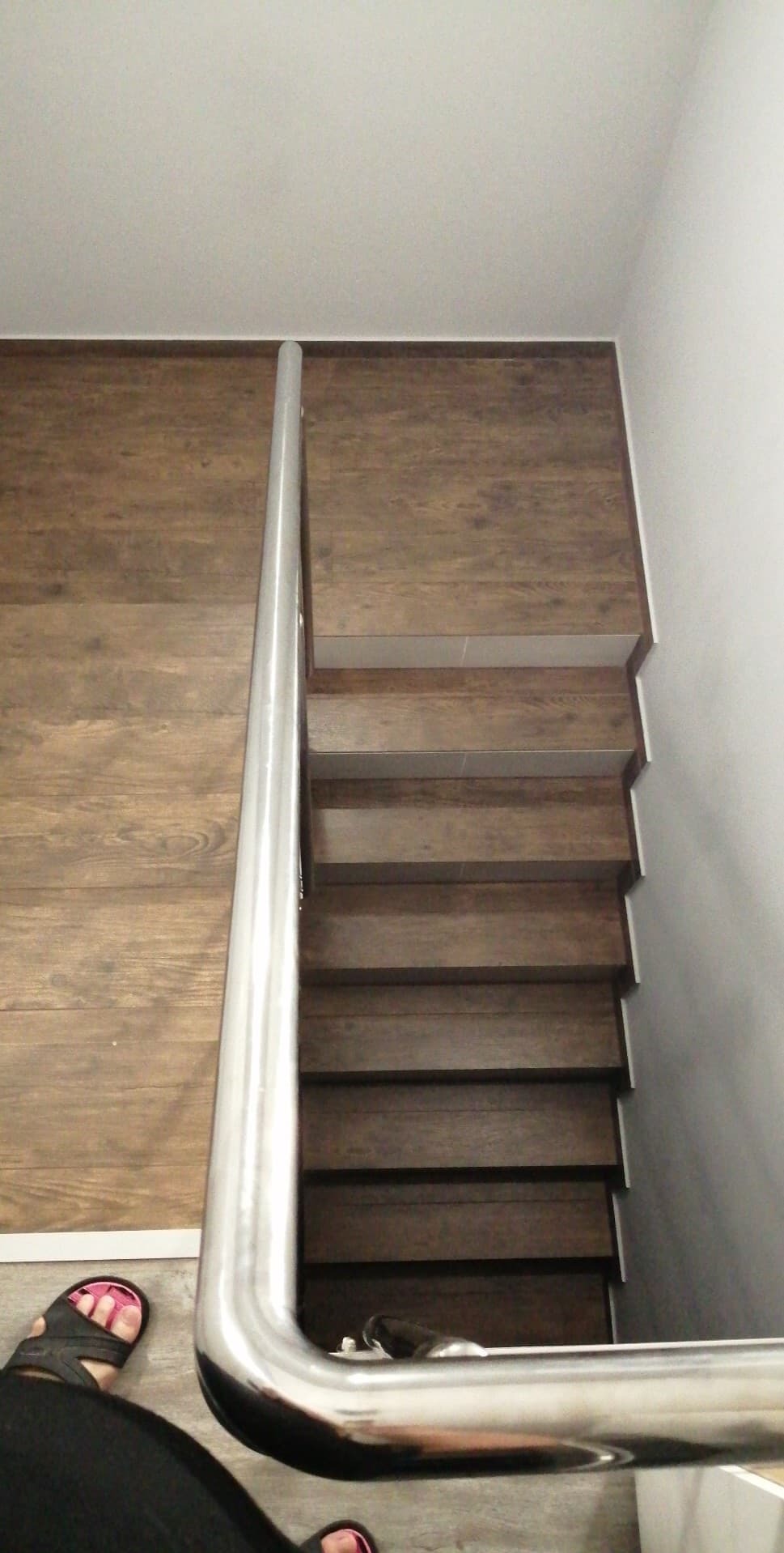 Schody wykończone panelami drewnopodobnymi z metalową poręczą, widok z góry. Fragment stopy w klapku w lewym dolnym rogu kadru. Minimalistyczne wykończenie.