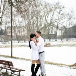 FotoOko - Andżelika Zychla - Zimowa sesja narzeczeńska w parku: mężczyzna całuje kobietę w czoło, oboje ubrani na biało, na tle ośnieżonego krajobrazu i ławek. Ujęcie pełne czułości i naturalności.
