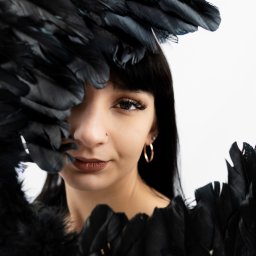 FotoOko - Andżelika Zychla - Portret kobiety z czarnymi piórami, złote kolczyki koła, piercing nosa, wyraziste rzęsy, ciemne włosy z grzywką na białym tle. Artystyczna sesja.