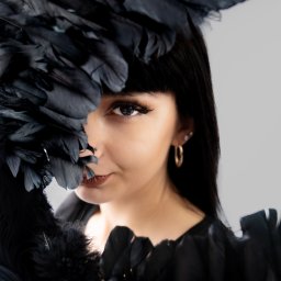 FotoOko - Andżelika Zychla - Portret kobiety z czarnymi piórami zasłaniającymi część twarzy, z widocznym piercingiem nosa i złotymi kolczykami na tle jasnoszarego studia.