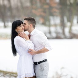 FotoOko - Andżelika Zychla - Zimowa sesja narzeczeńska: mężczyzna całuje kobietę w okularach, oboje ubrani na biało, na tle ośnieżonego parku z rozmytymi drzewami.