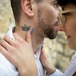 FotoOko - Andżelika Zychla - Profil zbliżający twarze pary: mężczyzna z tatuażem korony na szyi i obrączką, kobieta w okularach. Delikatne światło, kamienne tło.