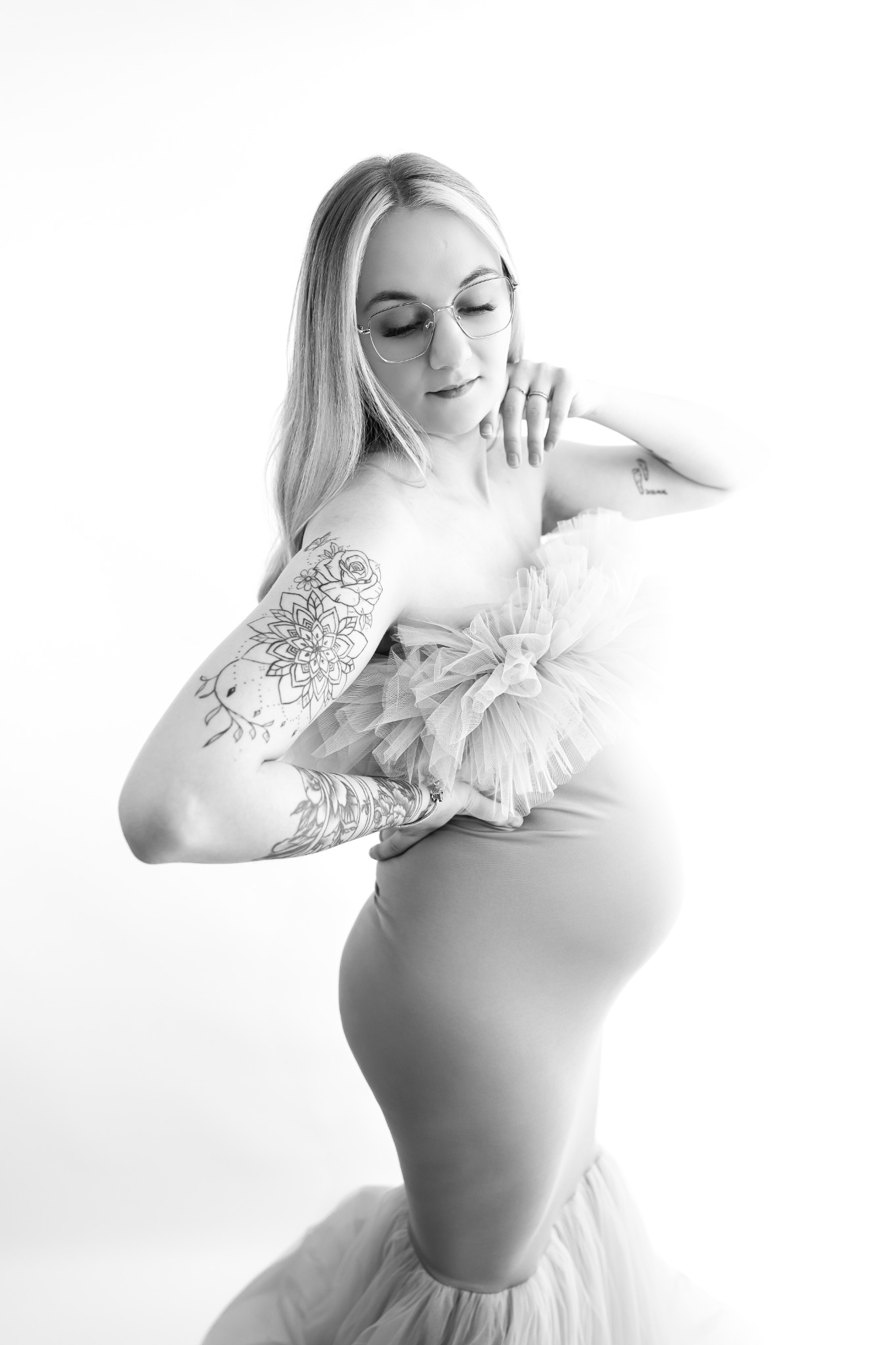 Monochromatyczne zdjęcie kobiety w ciąży, w sukni z tiulowym akcentem, z tatuażami na ramionach i okularach, pozującej na jasnym tle. Subtelne światło podkreśla kształty.