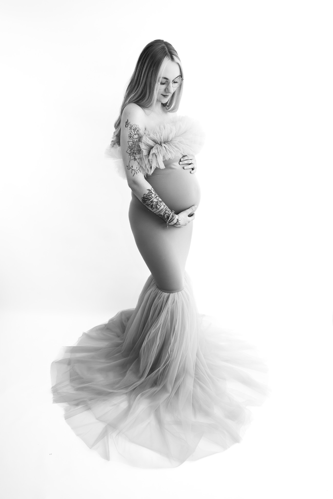 Czarno-białe zdjęcie kobiety w ciąży, w długiej sukni z tiulu, z tatuażami na ramionach, delikatnie spoglądającej w dół na swój brzuch, na jasnym tle.