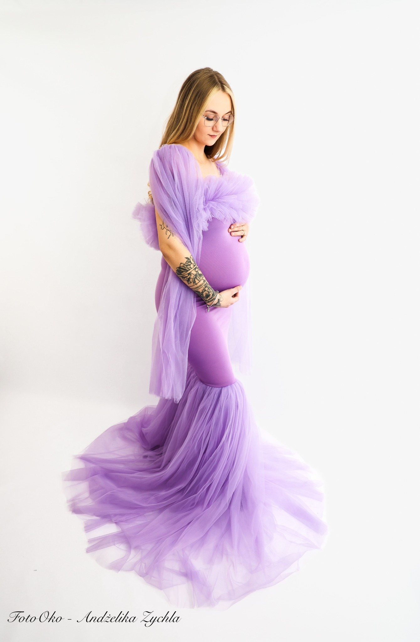 Kobieta w liliowej sukni ciążowej, z tatuażem na ramieniu, delikatnie spogląda w dół, trzymając ręce na brzuchu. Jasne tło podkreśla subtelność i intymność momentu.